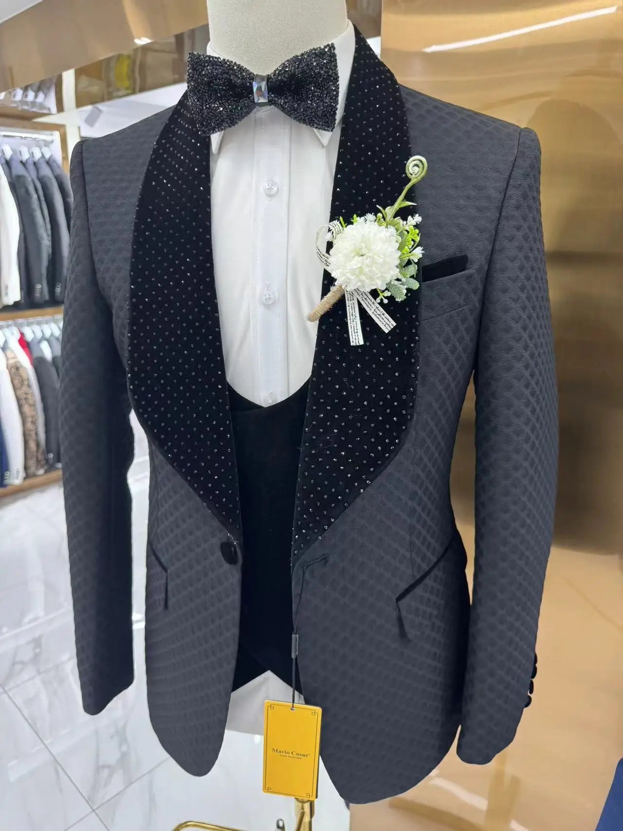 Gray textured suit with a black, studded shawl collar and matching bow tie, white shirt, and a boutonniere of white flowers. Displayed on a mannequin in a store.
