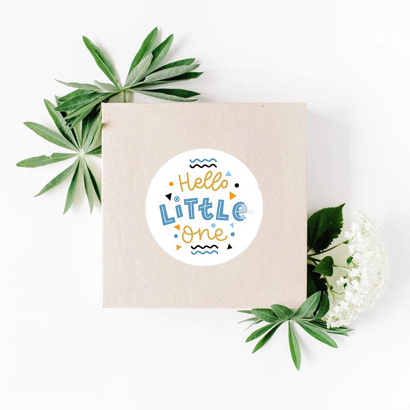 Brown gift box with a "Hello Little One" sticker in colorful text, surrounded by green leaves and small white flowers, set against a white background.