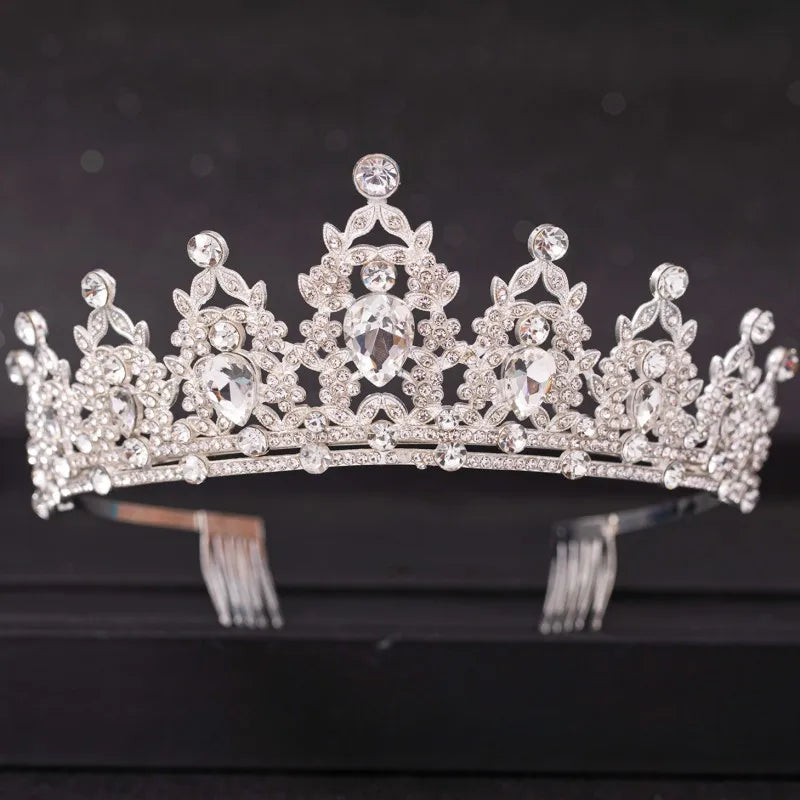 A sparkling silver tiara adorned with ornate patterns of clear gemstones and crystals, set against a dark background. The intricate design features teardrop and round gems.
