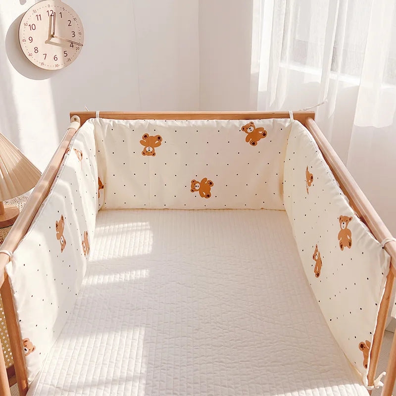 A cozy crib with soft beige bumpers featuring cute brown teddy bear patterns. The crib is set in a sunlit room with sheer curtains, a wall clock, and a table lamp nearby.