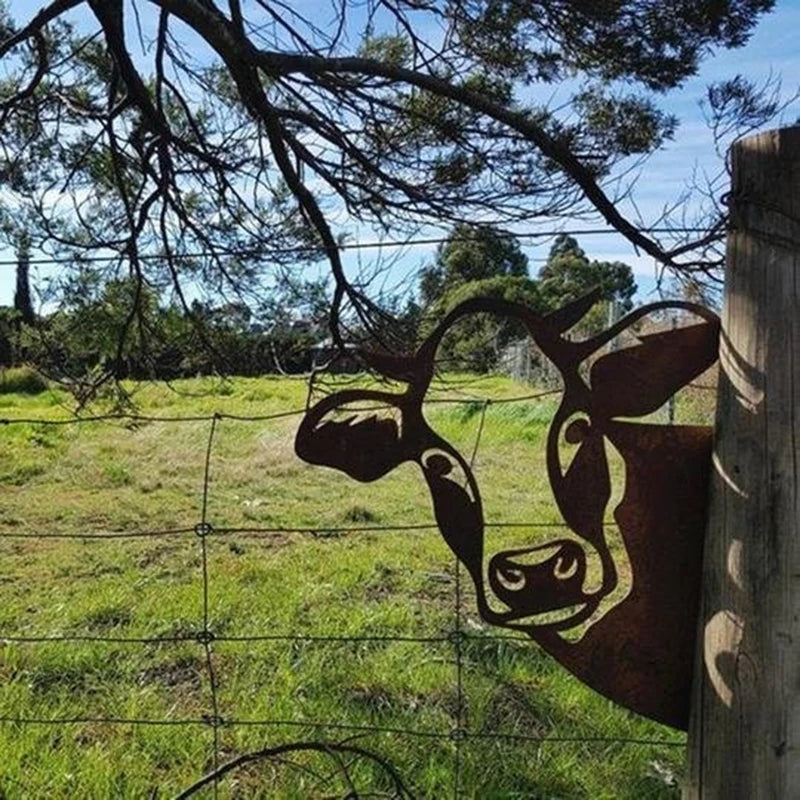 A rusty metal cow silhouette is fixed to a wooden post against a wire fence, creating a whimsical effect. In the background, bright green grass and trees are visible under a blue sky.