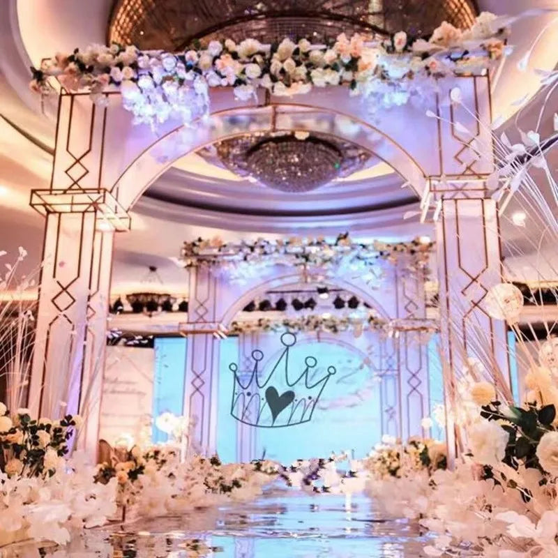 Elegant venue with arches adorned with white roses and soft pink lighting. A heart-topped crown is central on a reflective floor, under a grand chandelier.