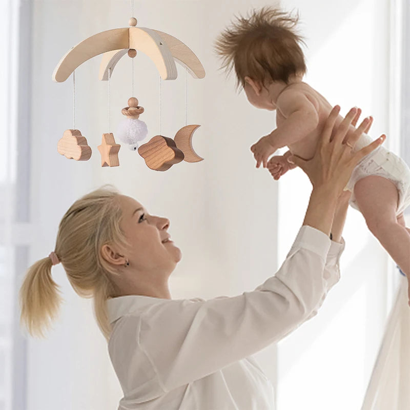 A woman in a white shirt joyfully lifts a baby in the air. Above them hangs a wooden mobile with star, cloud, moon, and pom-pom shapes, set in a softly lit room.