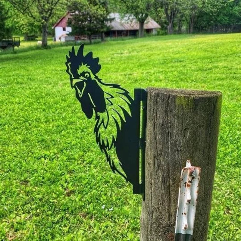 Metal silhouette of a rooster attached to a wooden post in a grassy field, with trees and a barn in the background.
