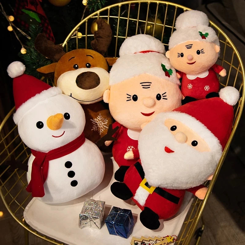 Plush toys of Santa, Mrs. Claus, a reindeer, and a snowman displayed on a chair with small wrapped gifts. Festive holiday decor is visible in the background.