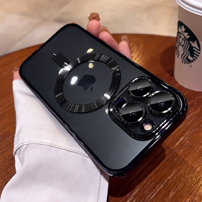 A person's hand holds a black iPhone with a MagSafe case featuring a ring design. The phone has three rear cameras and a Starbucks cup is visible on a wooden table in the background.