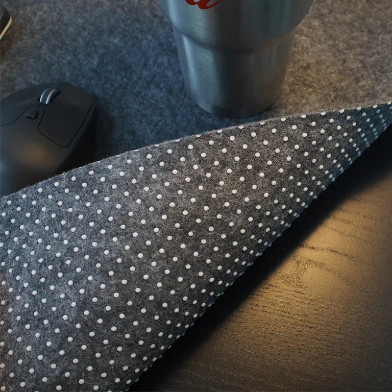 Close-up of a desk with a textured, non-slip mat partially rolled, a computer mouse, and a stainless steel tumbler. The mat has white polka dots and rests on a wooden surface.