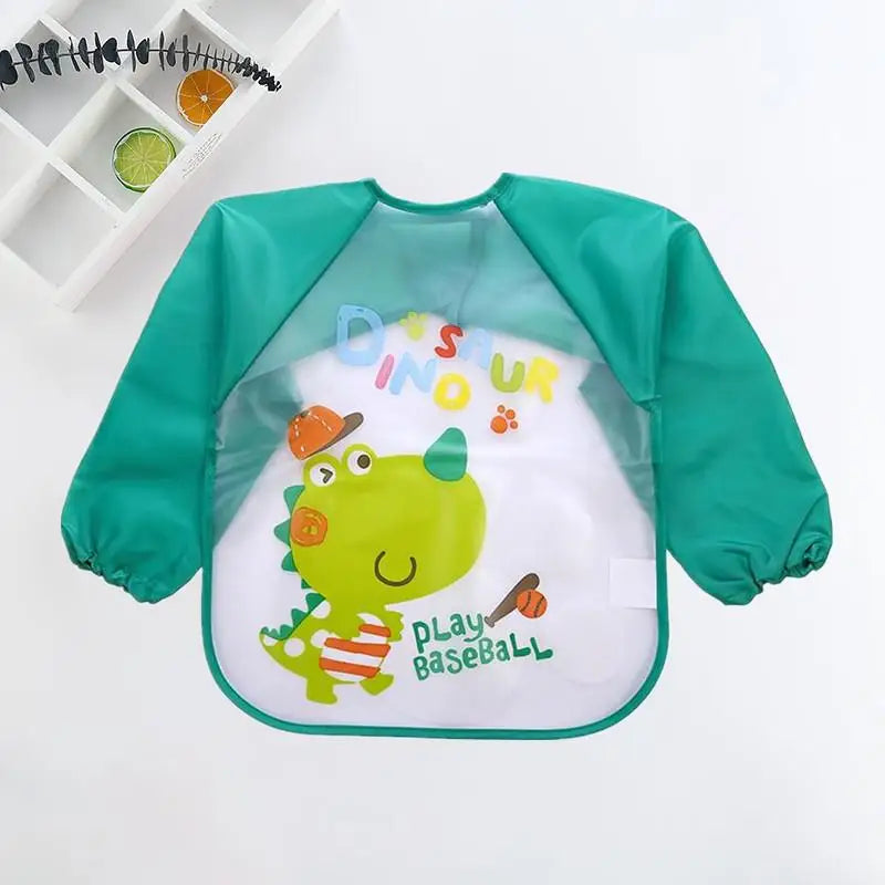 Green children's long-sleeve bib featuring a cute dinosaur with orange accents and the text "Play Baseball." A small shelf displays citrus slices in the background.