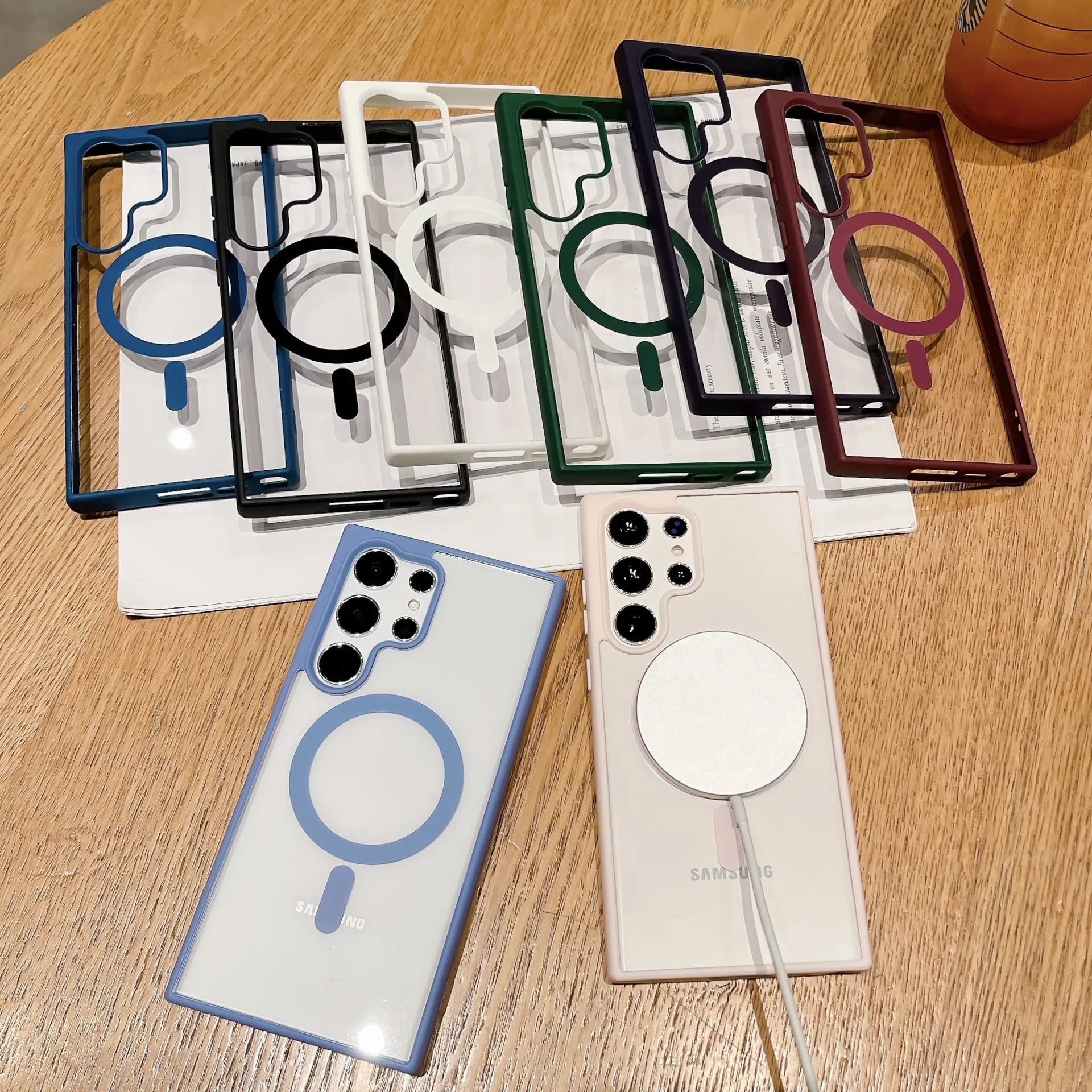 Assorted Samsung phone cases with magnetic circles are arranged on a wooden table, showcasing various colors like blue, white, green, and red. Two phones with cases are also visible.