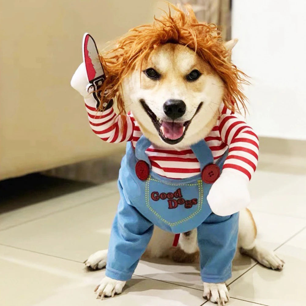 Dog dressed in a playful Chucky costume with red hair wig, striped shirt, overalls labeled "Good Dogs," and a fake knife. The dog is sitting indoors, looking cheerful.