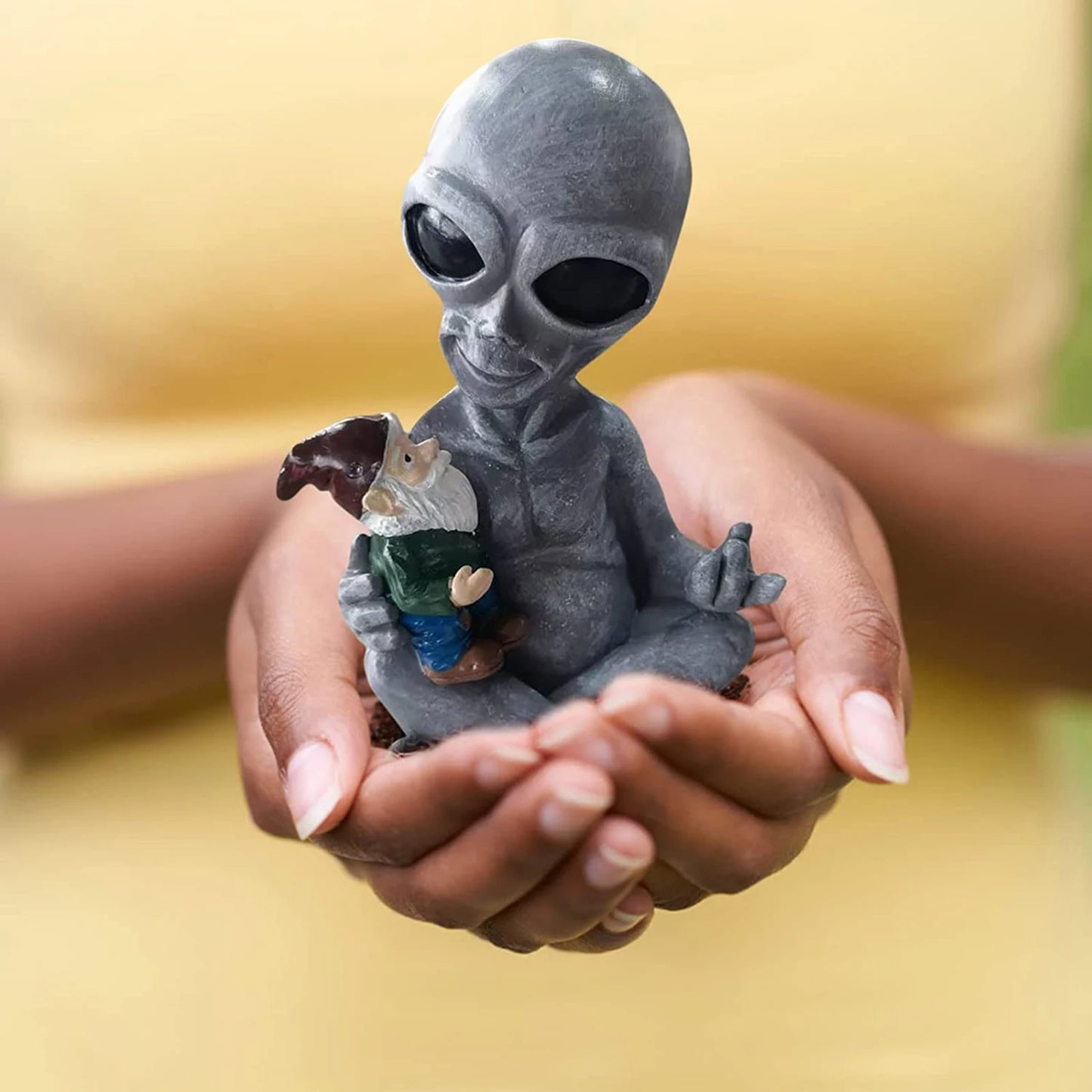Hands holding a statue of a gray alien seated cross-legged, gently cradling a small gnome, set against a background of a yellow shirt.