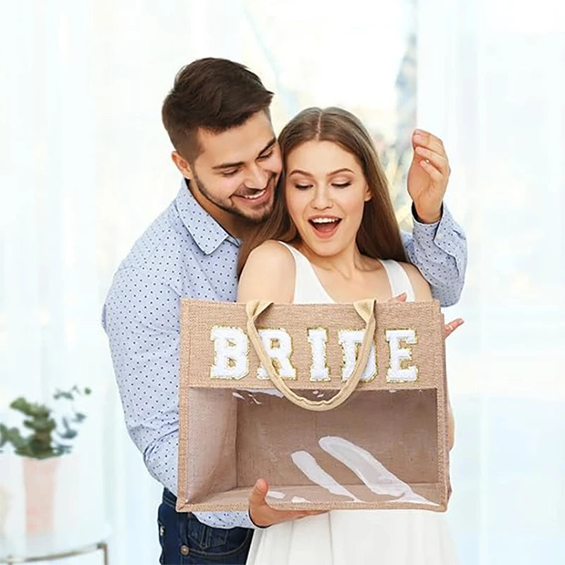 A smiling couple, with the woman surprised, holding a burlap tote bag labeled "BRIDE" in white letters. The man is hugging her from behind in a bright room.