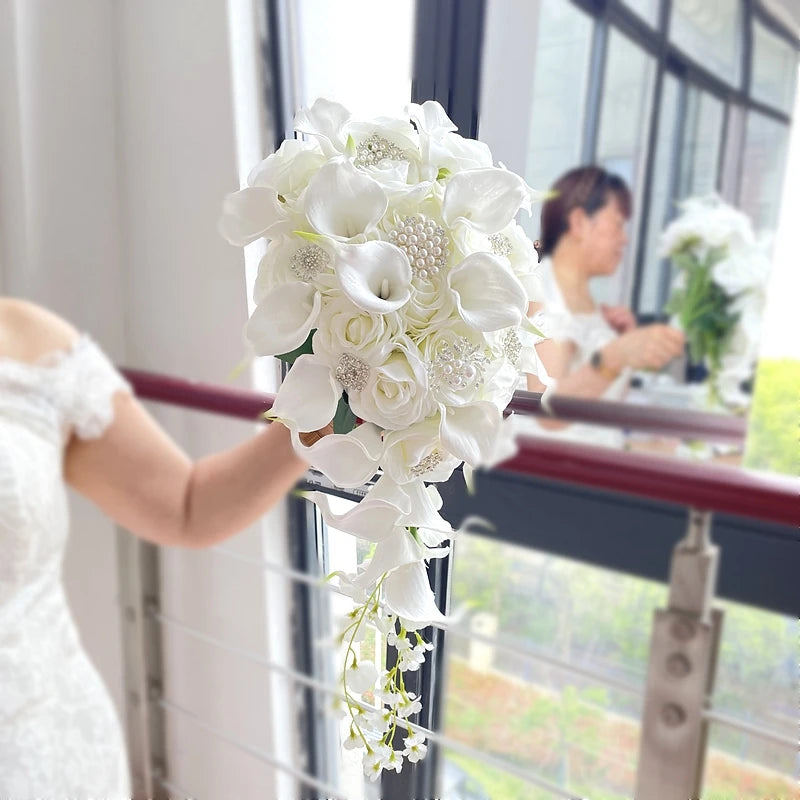 A person holds a cascading bouquet of white roses, calla lilies, and pearls in a bright room with large windows, reflecting a soft and elegant ambiance.