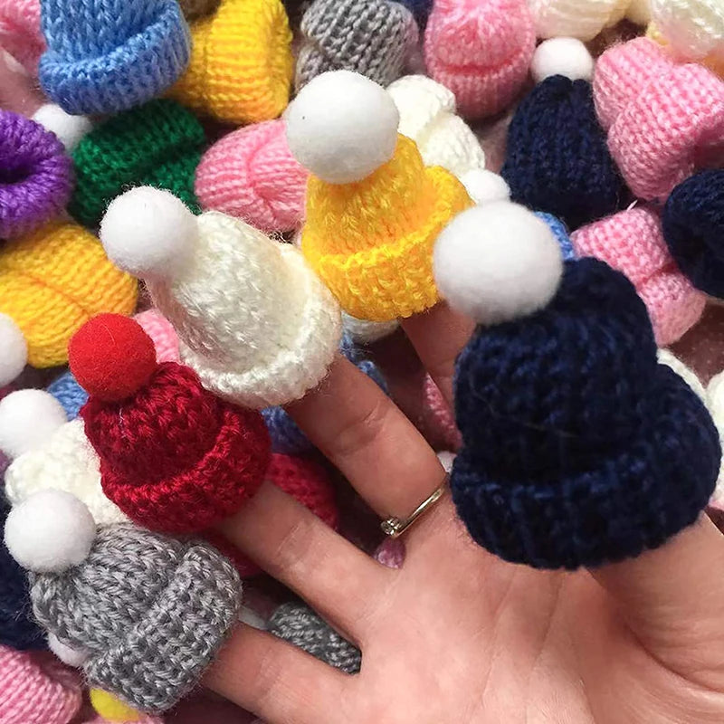A hand holds small knitted beanies in various colors, each with a pom-pom on top, surrounded by more mini beanies in different shades.