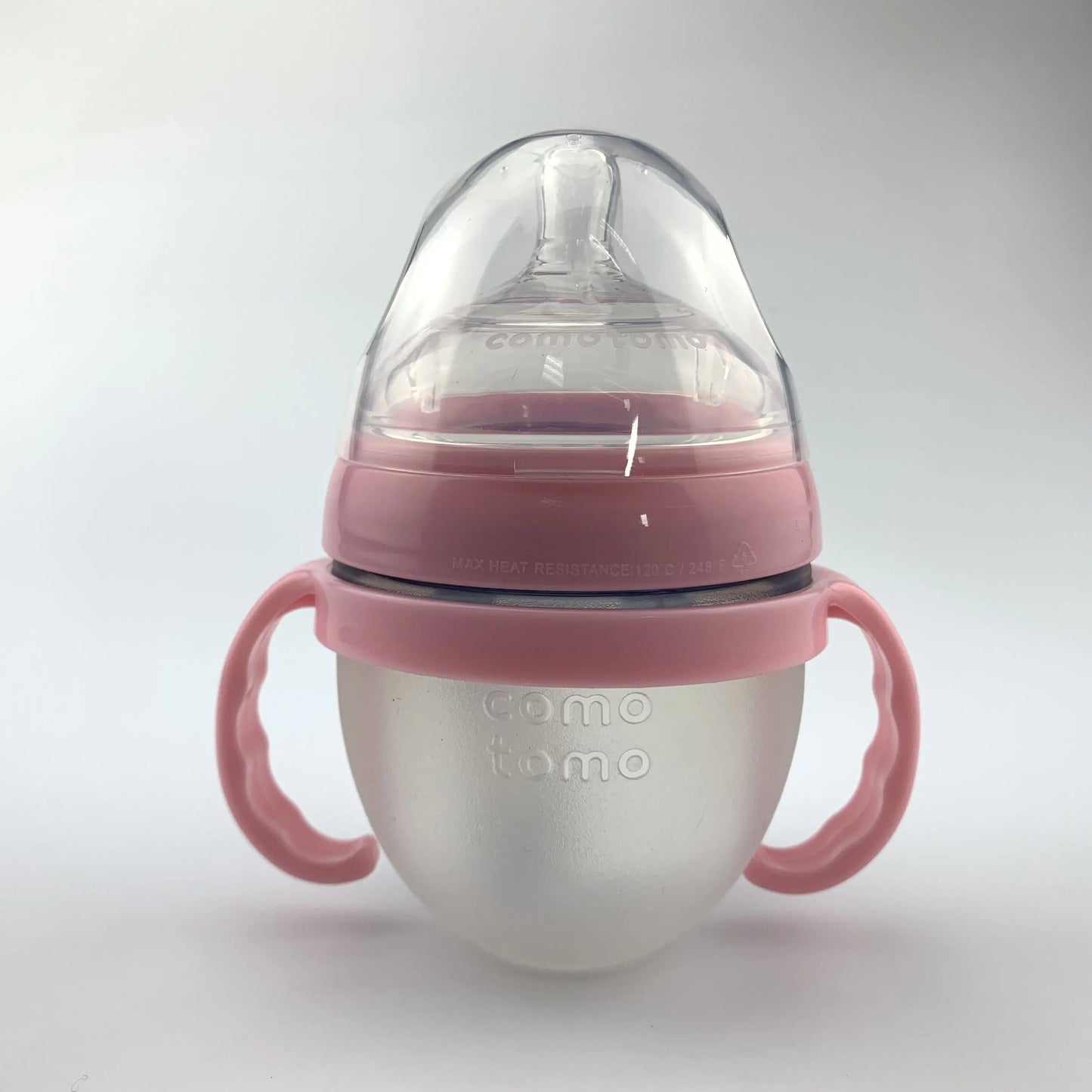 A baby bottle with a pink silicone base and handles, featuring a clear nipple and lid. The brand name "comotomo" is embossed on the frosted, rounded bottle surface.