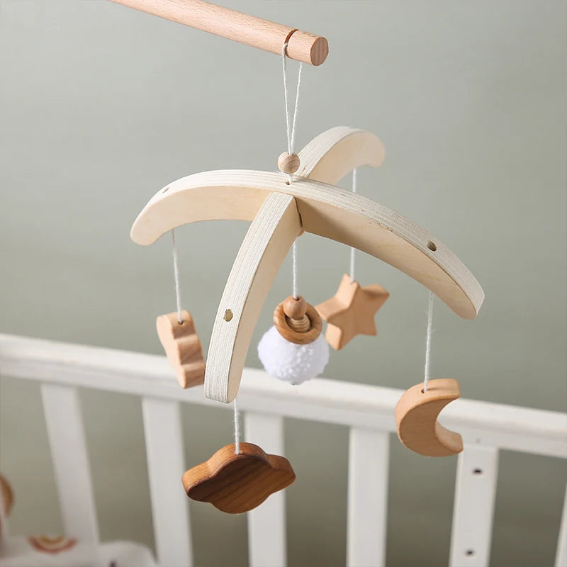 Wooden baby mobile with a star, cloud, moon, and pom-pom hanging from a crossbar, suspended over a white crib with a soft grey background.