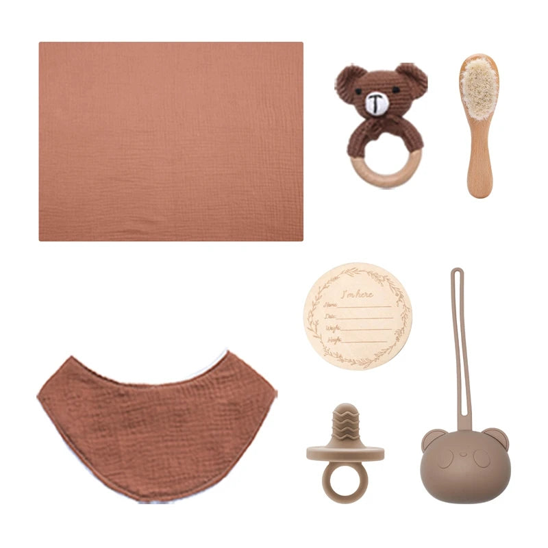 Baby essentials set including a brown blanket, bear-shaped teether, soft brush, bib, wooden name plaque, silicone pacifier, and a bear-shaped silicone case, arranged neatly.