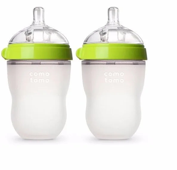 Two white baby bottles with silicone bodies and green lids, featuring a transparent nipple. The brand name, "comotomo," is printed on each bottle.