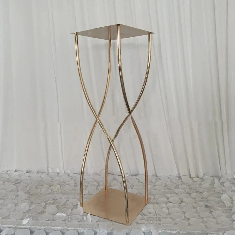 A tall, elegant gold metal pedestal with a modern, intertwining design stands on a textured white surface against a white curtain backdrop.