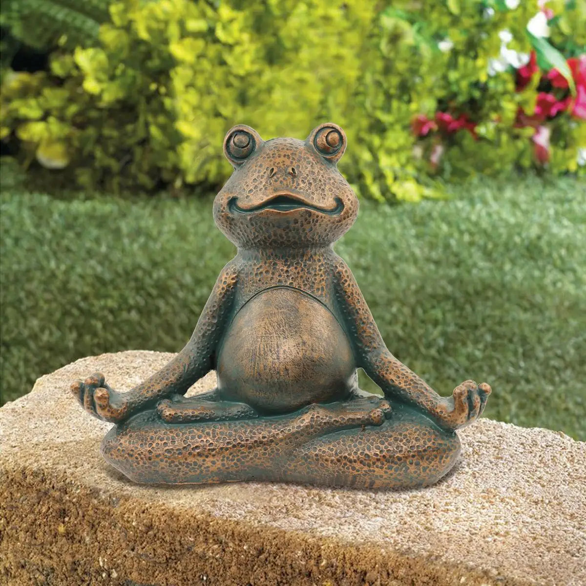 A frog statue in a yoga pose sits cross-legged on a rock. Its eyes are open, and it's smiling. The background features lush greenery and red flowers.