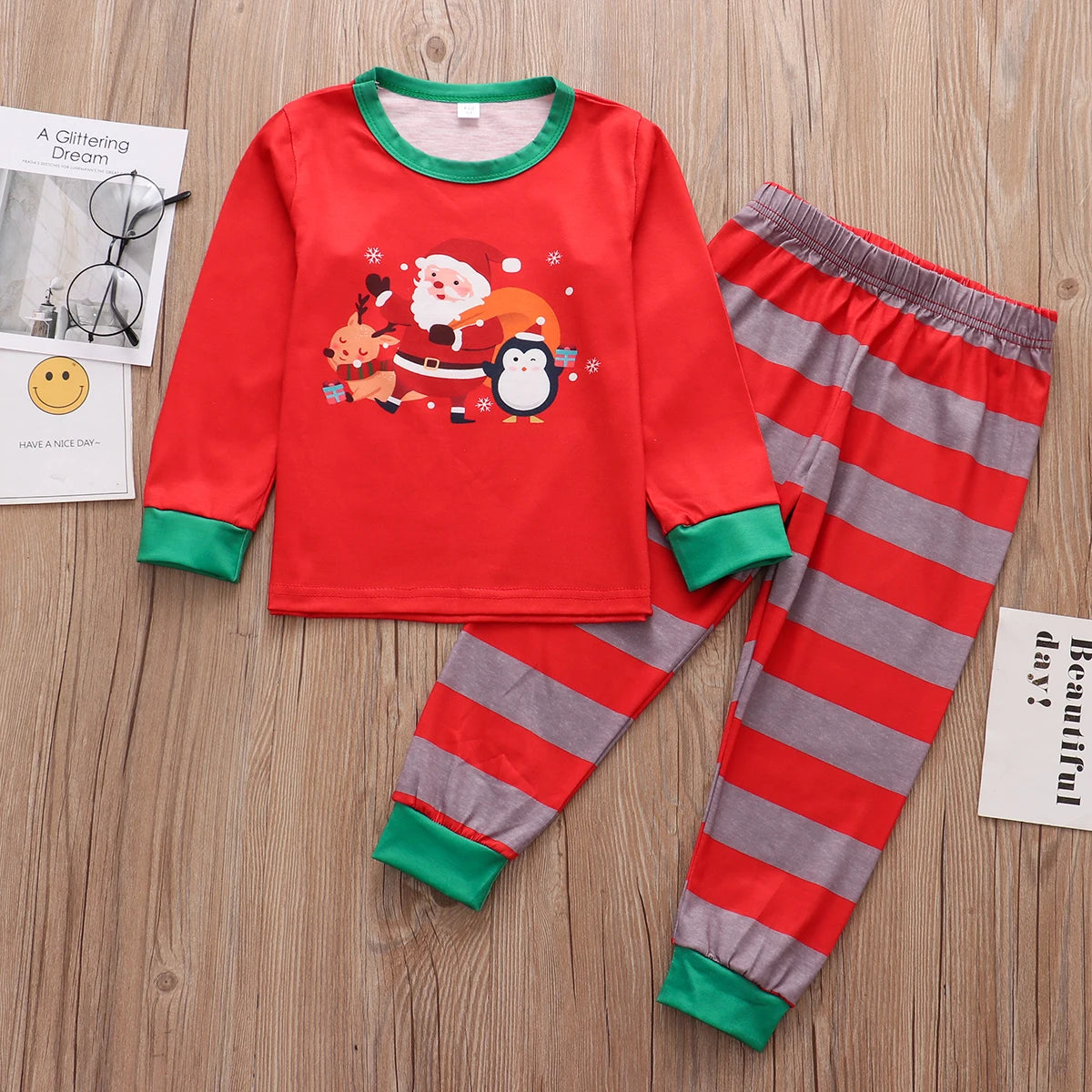 Red children's Christmas pajamas featuring Santa, a reindeer, and a penguin, with green cuffs. The pants are red and gray striped. Background includes glasses and papers on wooden floor.
