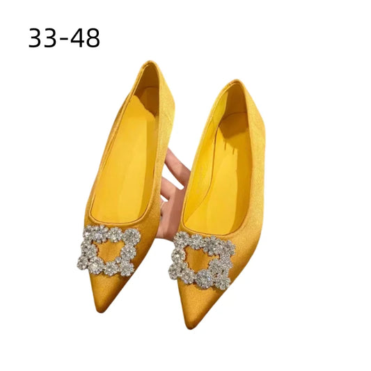 Yellow pointed-toe flats embellished with sparkling crystal buckles. A hand holds the shoes against a white background. The text "33–48" indicates the size range.