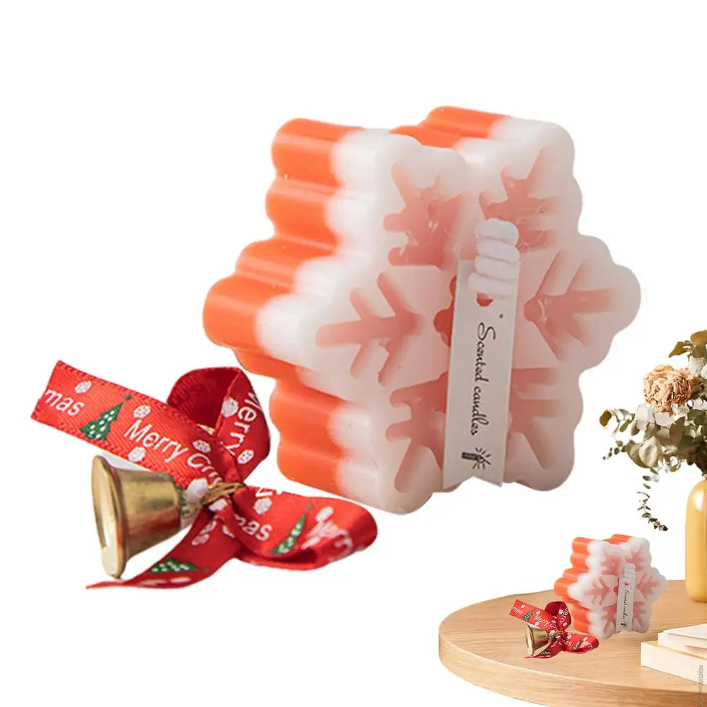 Snowflake-shaped orange and white scented candle with a "Merry Christmas" ribbon and bell. Displayed on a wooden surface next to a vase with flowers.