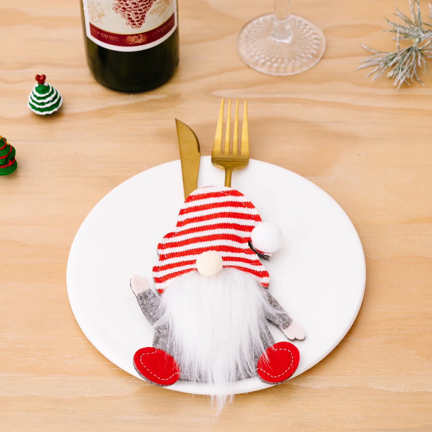 Festive table setting with a gnome cutlery holder on a plate, beside a bottle of wine, a glass, and mini Christmas trees on a wooden surface.