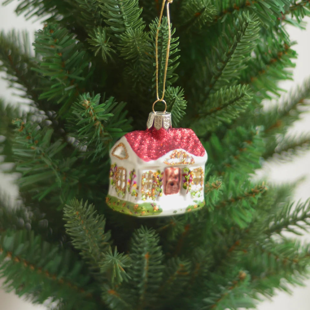 A festive house-shaped ornament with glittery red roof hangs on a lush green Christmas tree, symbolizing holiday cheer and decoration.