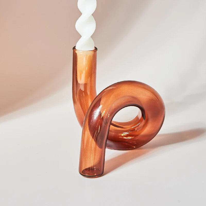 Twisted white candle in a looping, amber glass holder with a smooth, curvy design, showcased on a light beige background.