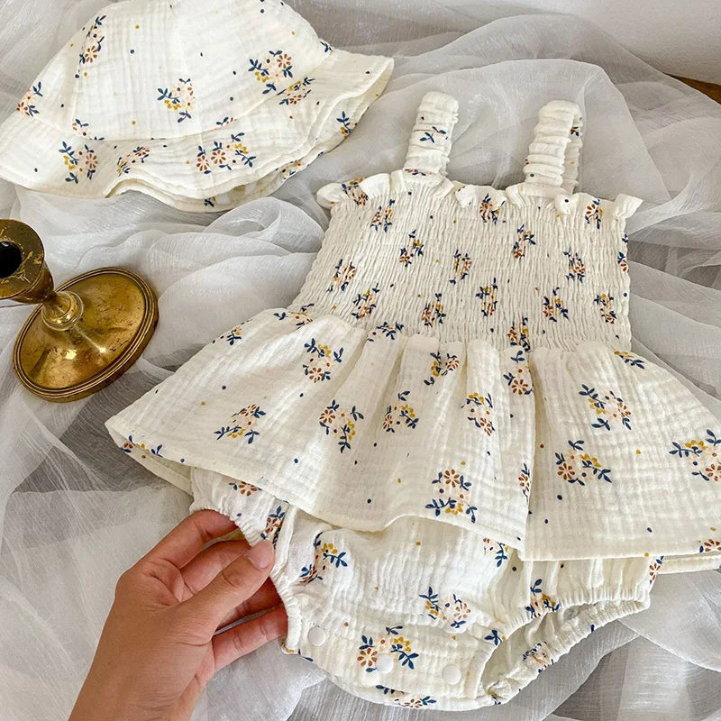 Baby outfit with floral print on white fabric, featuring a dress with straps, matching bloomers, and a hat, displayed on a white sheer cloth beside a small brass candlestick.
