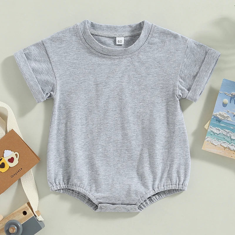 A light gray baby romper with short sleeves and snap buttons at the bottom, surrounded by a toy camera, postcard, and a card with coffee cup illustrations on a beige background.