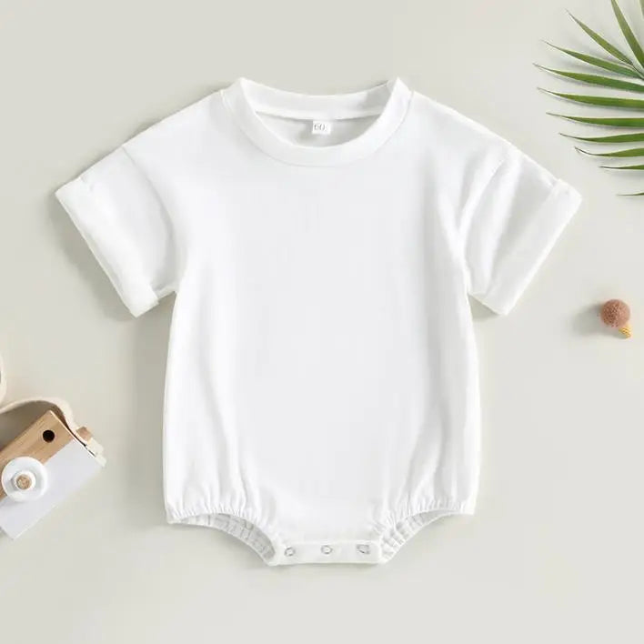 White baby onesie with short sleeves and snap buttons at the bottom. It is placed on a neutral background with a wooden toy and small plant on the side.