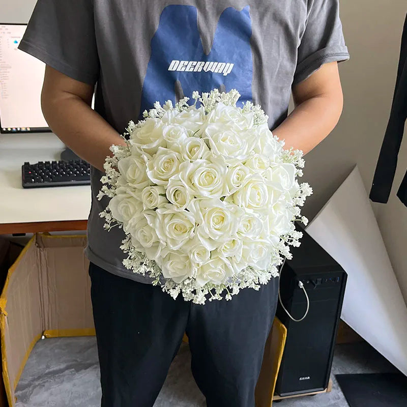 Person holding a bouquet of white roses and baby's breath, standing indoors. The person is wearing a gray shirt with a blue and white design. A desk with a keyboard is visible behind.