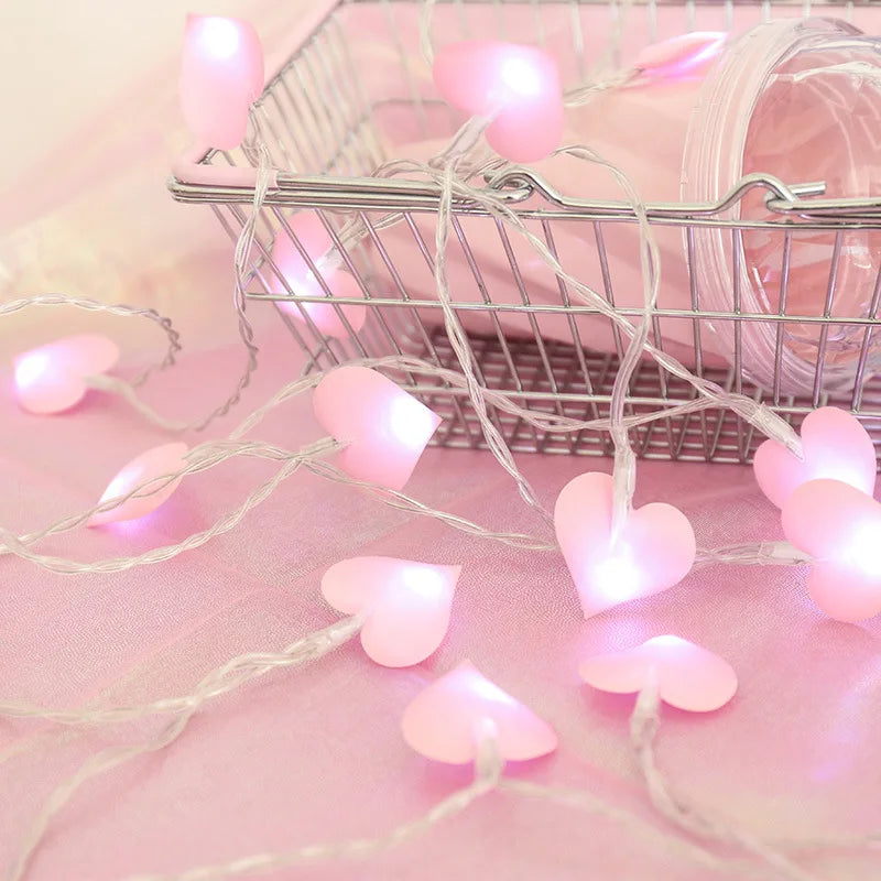 Pink heart-shaped string lights draped over a wire basket on a soft pink surface, creating a warm and romantic ambiance.