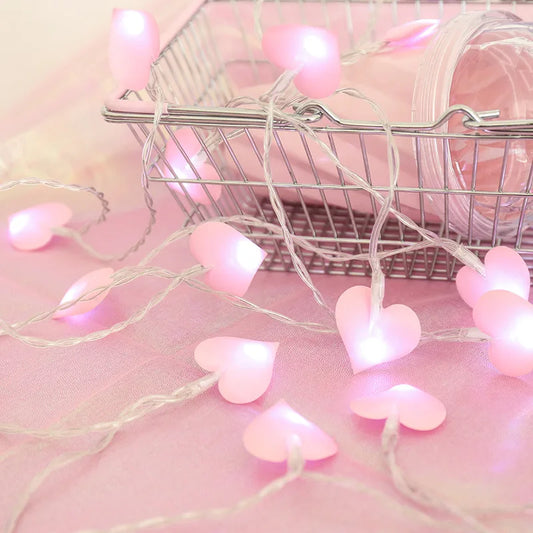 Pink heart-shaped string lights draped over a wire basket on a soft pink surface, creating a warm and romantic ambiance.