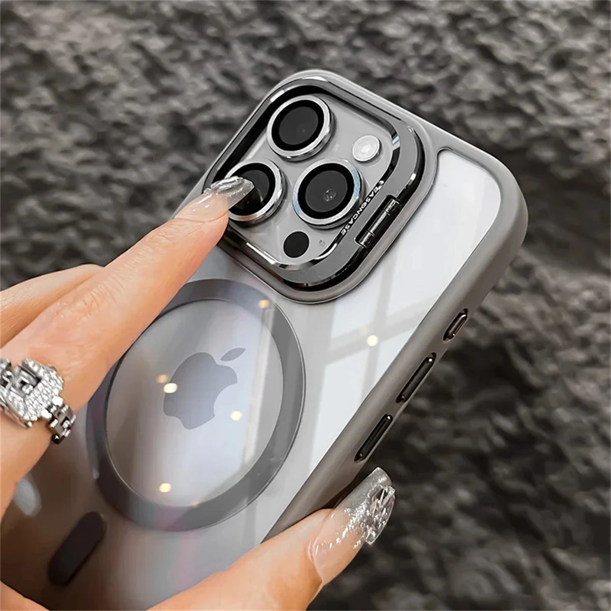 Close-up of a hand with shimmering nails pointing at the camera lens of a smartphone in a clear case, featuring an Apple logo. The background is a textured gray surface.