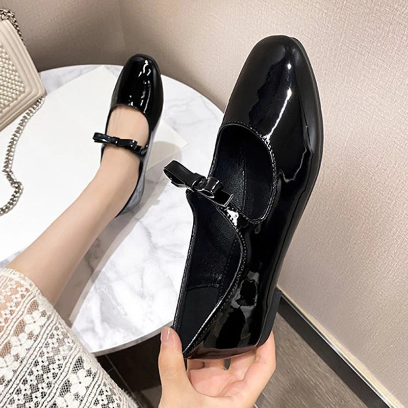 A pair of shiny black patent leather Mary Jane shoes with bows, one held in hand, the other worn. The wearer sits on a white marble table next to a beaded handbag.