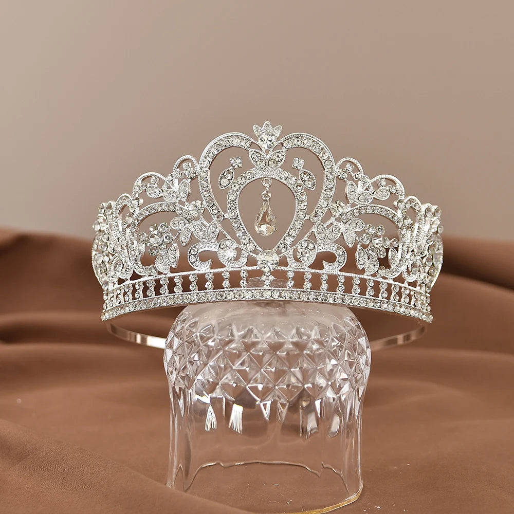 Ornate silver tiara adorned with sparkling gemstones, featuring heart-shaped designs and a dangling teardrop gem, resting on a textured glass stand with a brown backdrop.