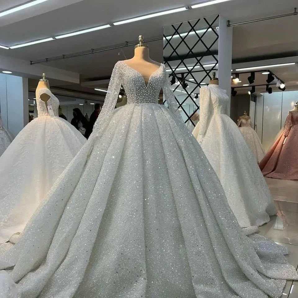 A sparkling wedding gown on a mannequin features a deep V-neck and full skirt, surrounded by other elegant white and pink gowns in a modern showroom setting.