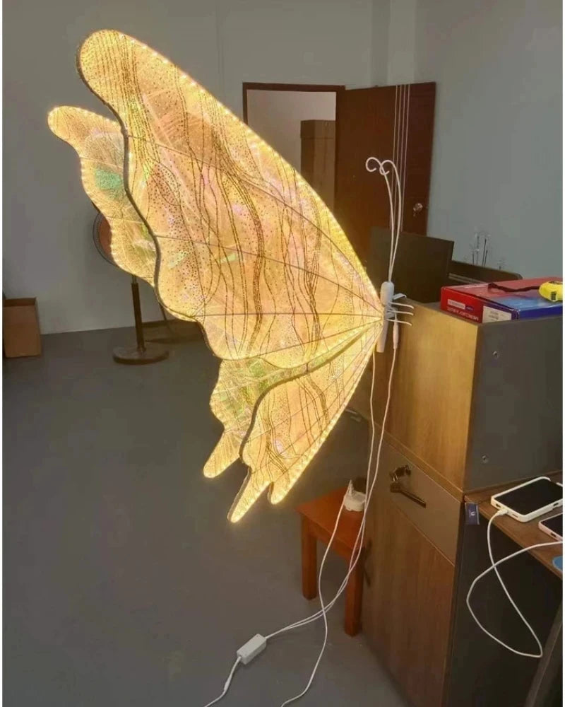 A large, illuminated butterfly wing lamp is attached to a wooden cabinet in a room. The translucent wing glows with warm, yellow light, revealing intricate patterns and veins.