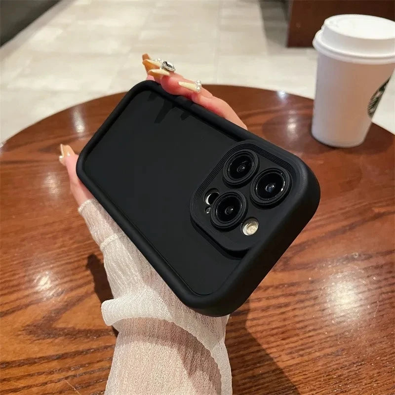 A hand holding a black smartphone with a triple camera in a café setting; a white to-go cup is on the wooden table in the background.