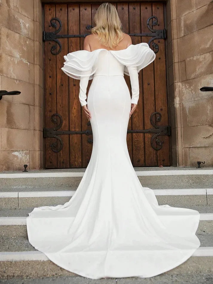 A woman in a white off-the-shoulder gown with dramatic ruffled sleeves stands on stone steps, facing an ornate wooden door. The dress has a long, flowing train.