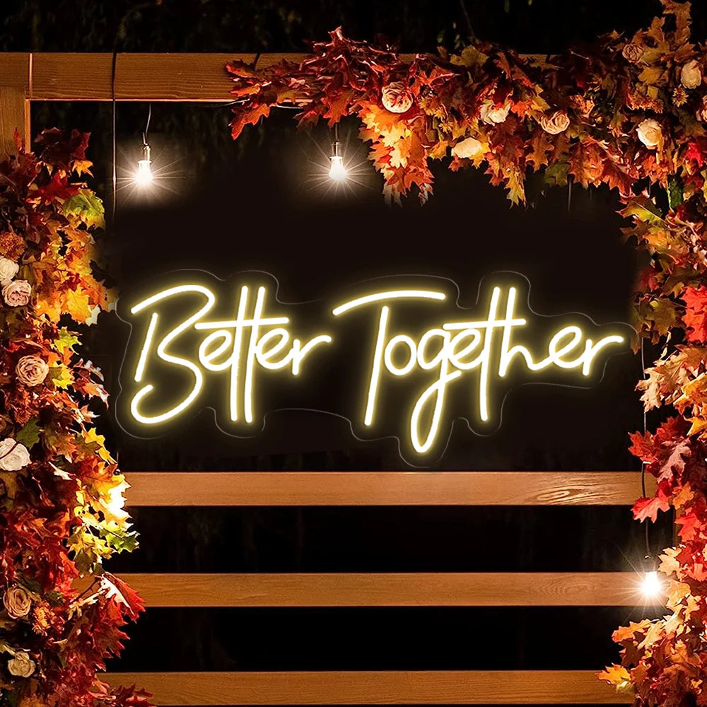 Neon sign reading "Better Together" surrounded by autumn leaves and flowers on a wooden frame, illuminated by small hanging lights.