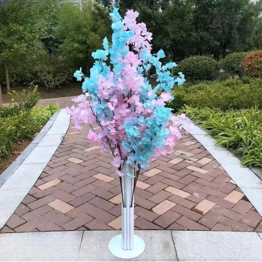 Artificial tree with pink and blue leaves stands on a paved path in a garden, surrounded by various green foliage and trees in the background.