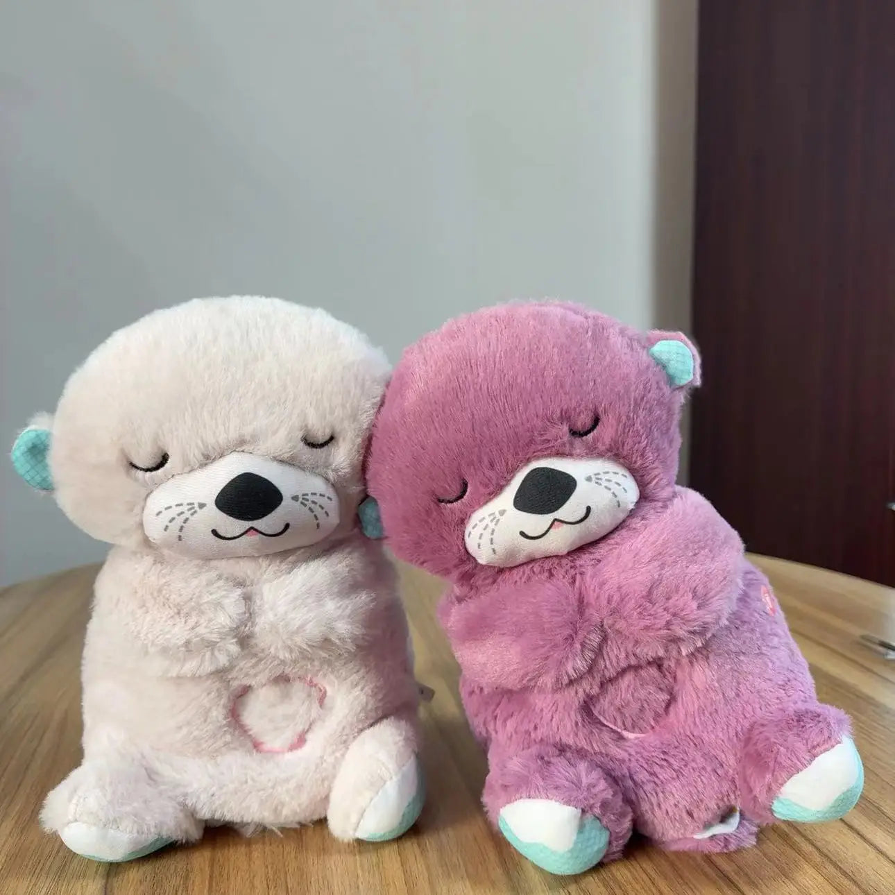 Two plush otter toys with eyes closed, one white and the other purple, sit side by side on a wooden table. Both have embroidered hearts on their chests and teal ears and feet.