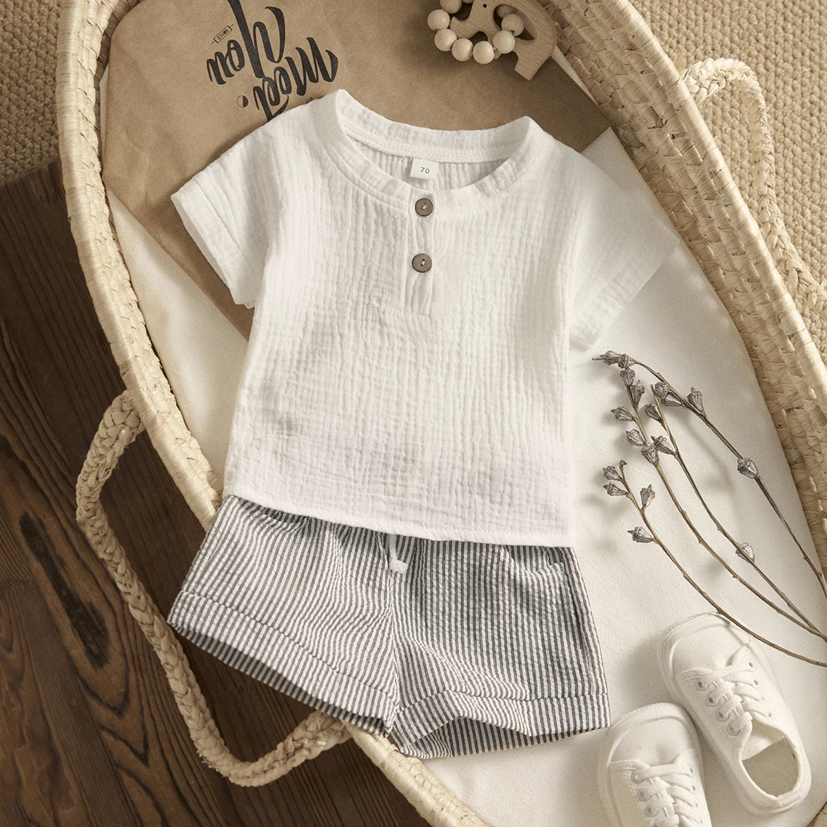 A baby outfit featuring a white buttoned shirt and striped shorts, placed on a woven basket with decorative twigs and white shoes on a wooden floor.