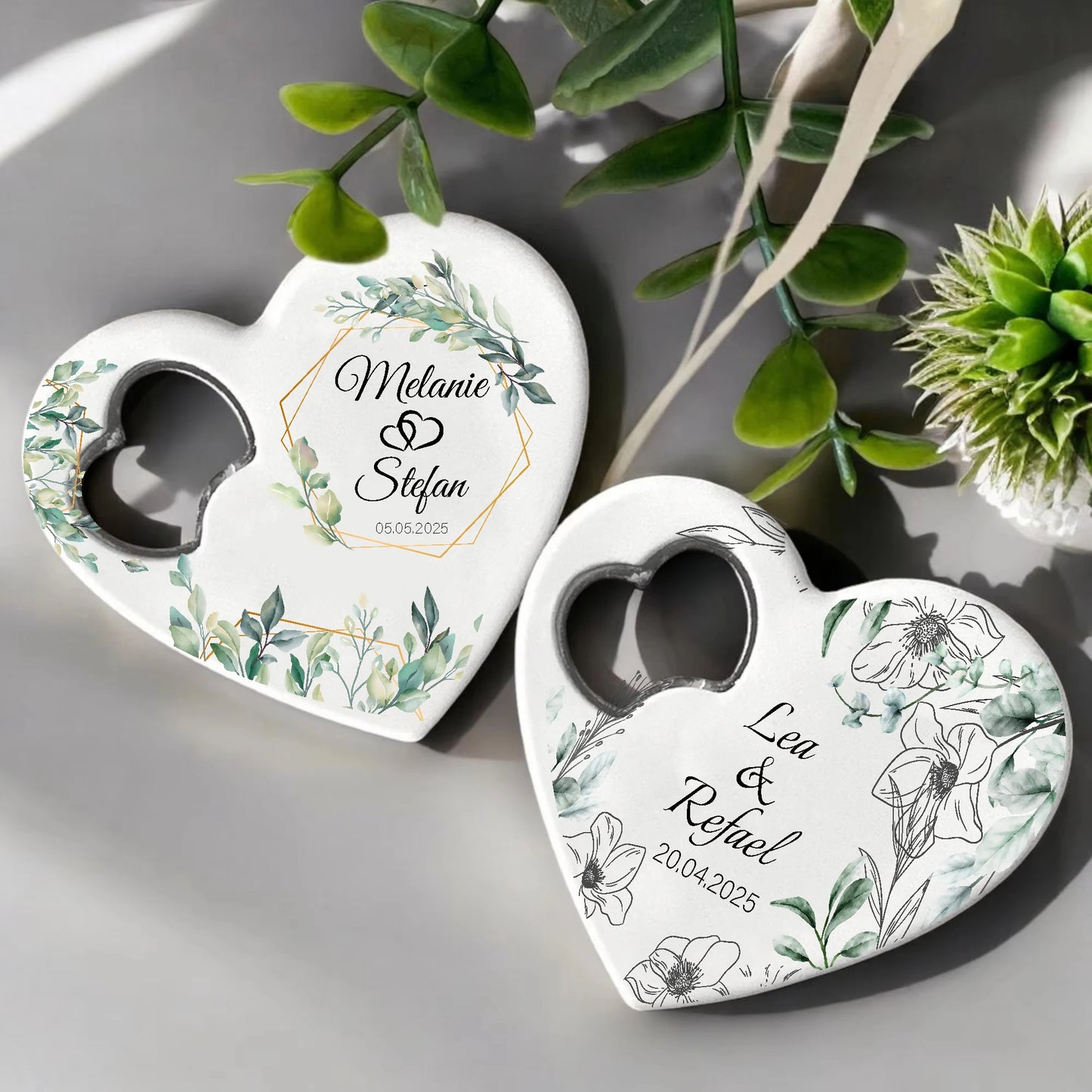 Two heart-shaped objects with floral designs and names inscribed: "Melanie & Stefan, 05.05.2025" and "Lea & Rafael, 20.04.2025," surrounded by green leaves.