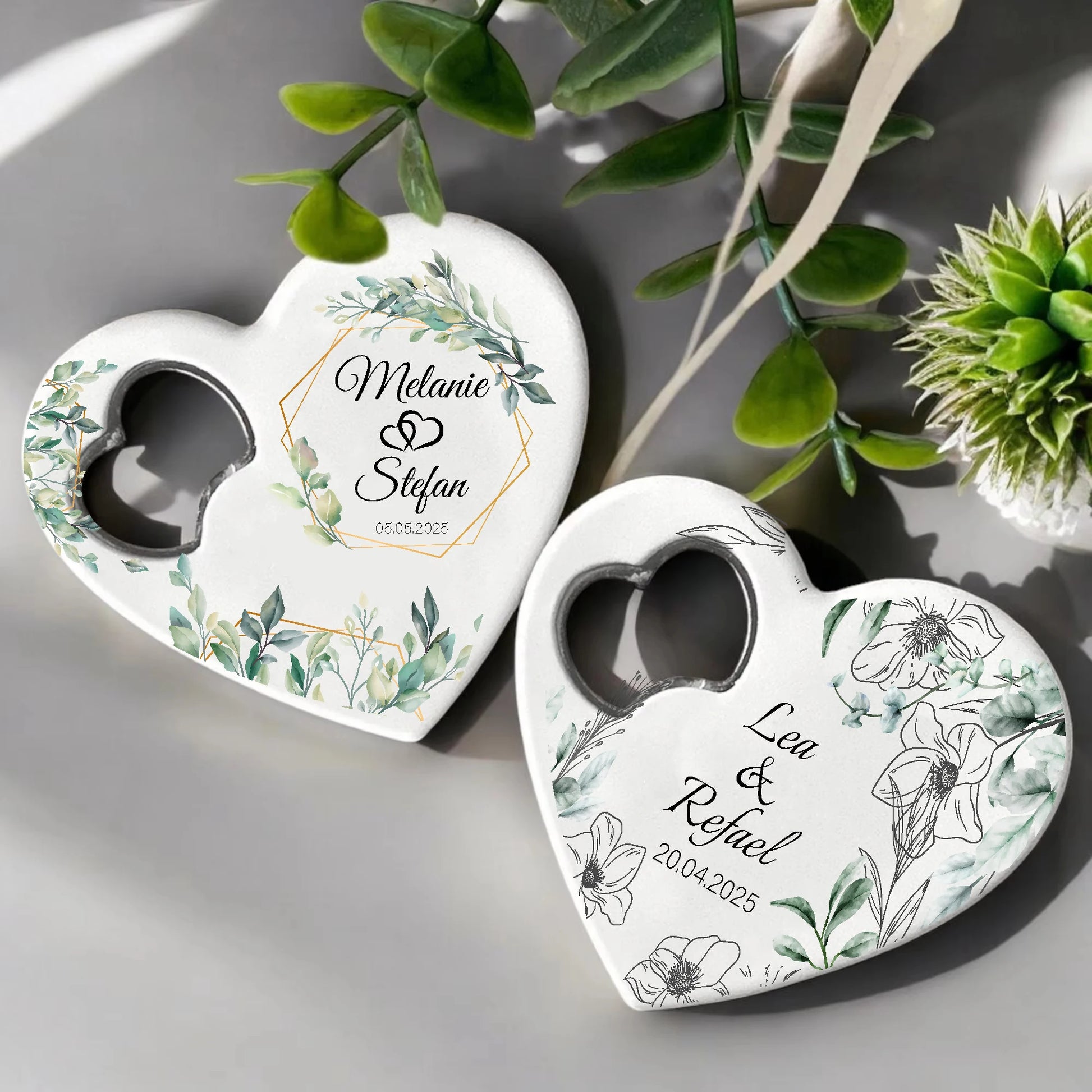 Two heart-shaped objects with floral designs and names inscribed: "Melanie & Stefan, 05.05.2025" and "Lea & Rafael, 20.04.2025," surrounded by green leaves.