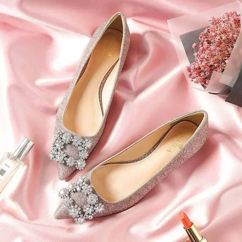 Elegant pink glitter flats with jeweled embellishments on pointed toes, placed on silky fabric. Nearby are a bouquet of pink flowers, lipstick, and perfume.